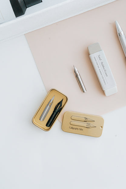 Gold tin with calligraphy nibs, a loose nib, a sand eraser, and a silver mechanical pencil on a white and blush background