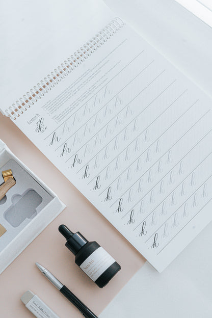 Spiral-bound calligraphy practice book open to the letter 'h,' with sumi ink, a mechanical pencil, and a sand eraser on a desk
