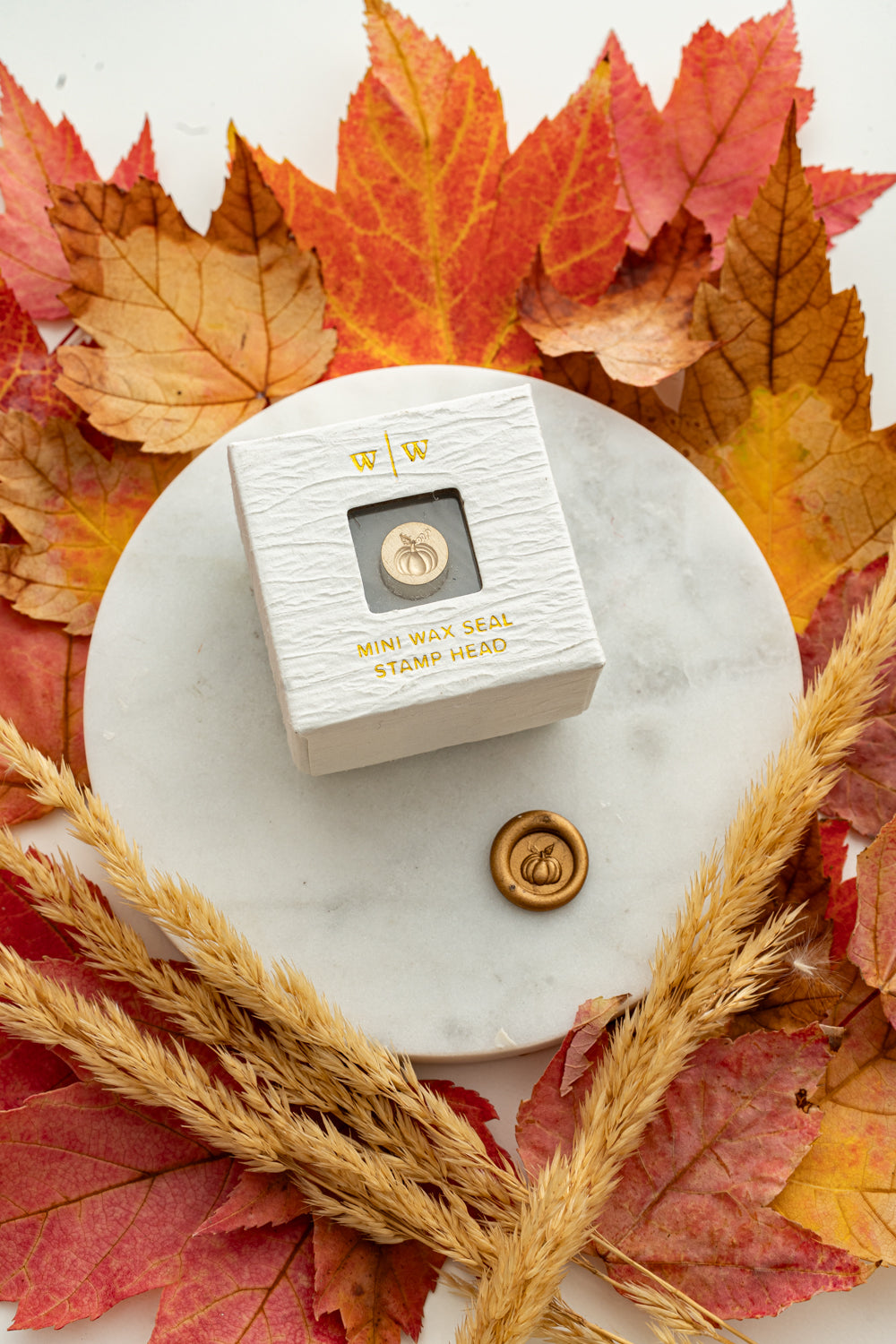 Mini pumpkin wax seal stamp head in white packaging with gold foil lettering, paired with a gold wax seal impression on a marble surface surrounded by autumn leaves and wheat stems.