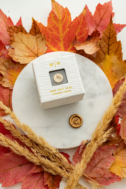 Mini pumpkin wax seal stamp head in white packaging with gold foil lettering, paired with a gold wax seal impression on a marble surface surrounded by autumn leaves and wheat stems.