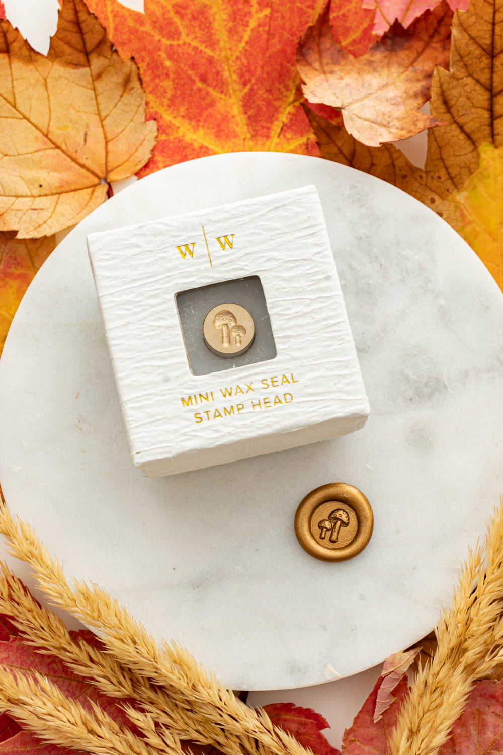 Mini mushroom wax seal stamp head in white packaging with gold foil lettering, paired with a gold wax seal impression on a marble surface surrounded by cozy fall leaves and wheat stems.