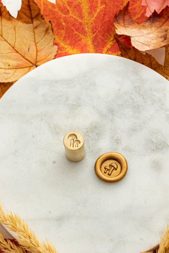 Gold mini wax seal stamp engraved with a pair of mushrooms, shown beside its wax impression on a marble surface surrounded by autumn leaves and golden wheat stems.