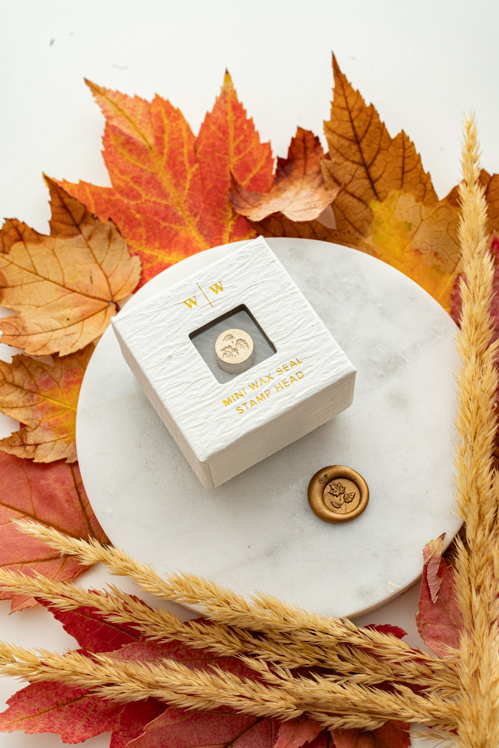 Mini fall leaves wax seal stamp head in white packaging with gold foil lettering, paired with a gold wax seal impression on a marble surface surrounded by autumn leaves and wheat stems.