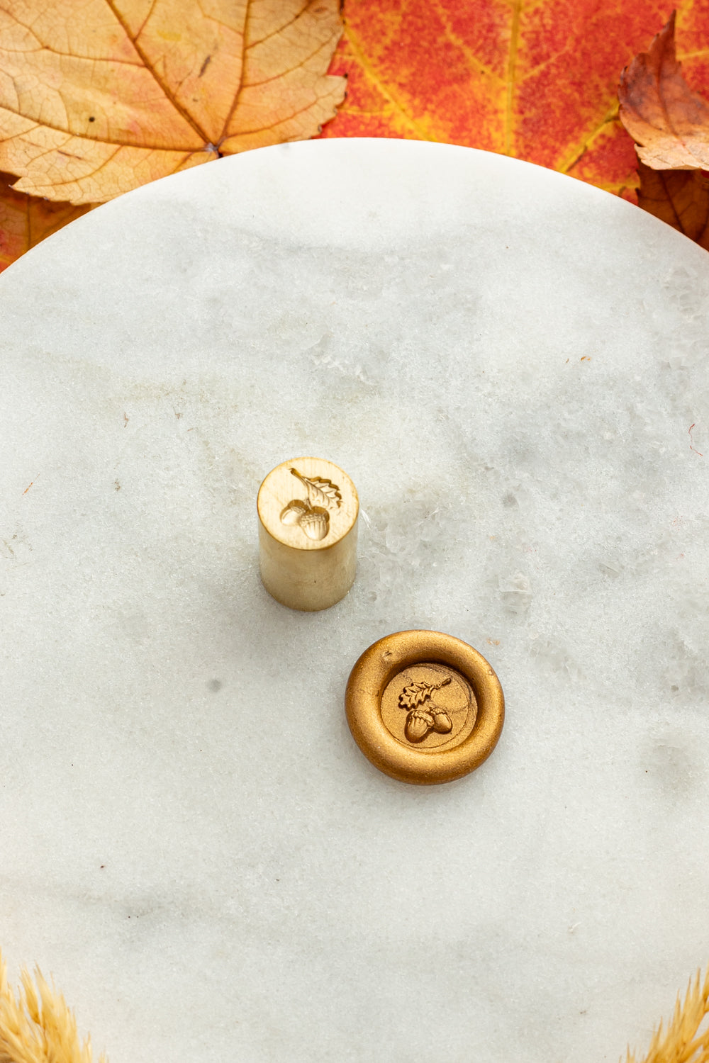 Gold mini wax seal stamp engraved with an acorn, shown beside its wax impression on a marble surface surrounded by autumn leaves and golden wheat stems.
