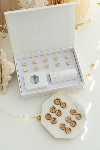 Holiday Mini Wax Seal Head Set opened to reveal twelve brass stamp designs, paired with their gold wax seal impressions on a marble slab, surrounded by soft festive décor and string lights.