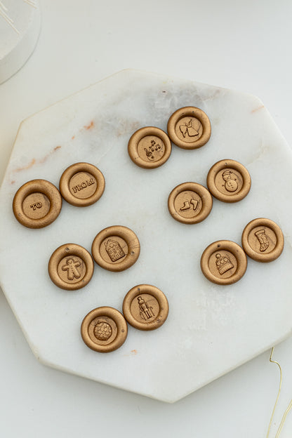 Flat lay of gold wax seals showcasing all twelve Holiday Mini Wax Seal Stamp designs, including festive icons like a snowman, nutcracker, stocking, and “TO” and “FROM” imprints, displayed on a marble slab.