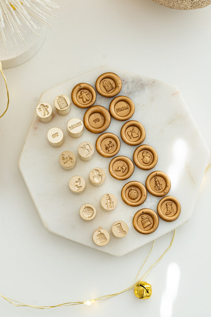 Flat lay of the Holiday Mini Wax Seal Head Set showing all twelve festive brass stamp designs and their matching wax seal impressions on a marble slab with gold holiday lights.