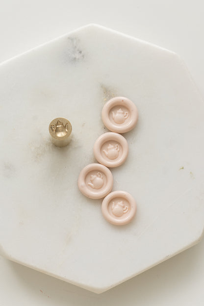 A mini brass wax seal stamp head with a 3D tea pot design beside a bunch of blush mini wax seals on a white marble coaster