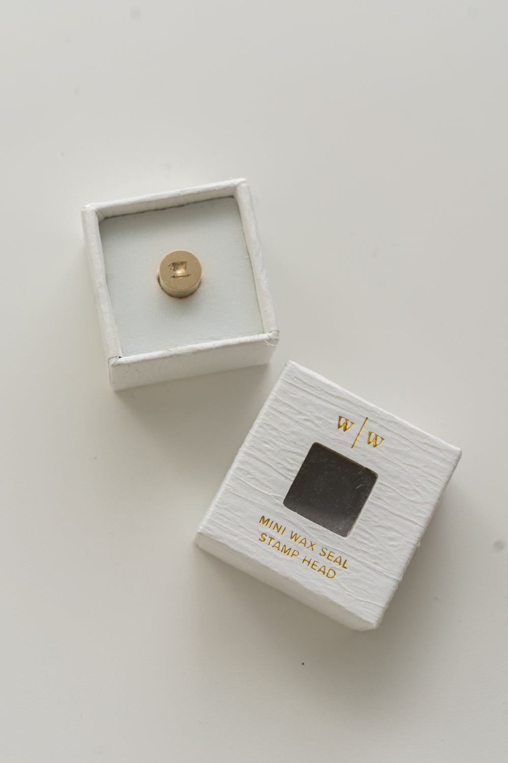A mini brass wax seal stamp head with a 3D tea cup design in a textured white ring-style box, with its lid beside it