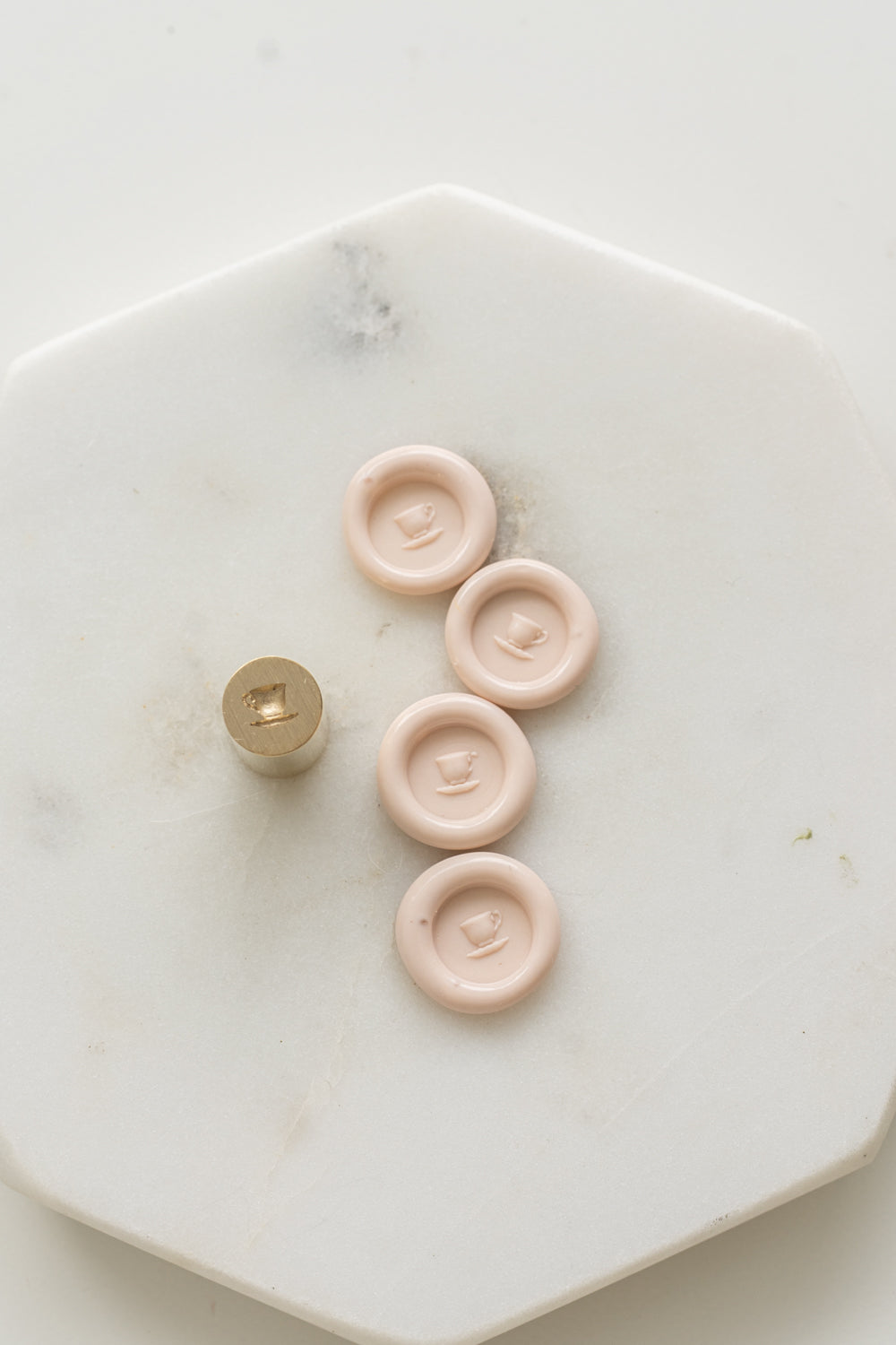 ​​A mini brass wax seal stamp head with a 3D tea cup design beside a bunch of blush mini wax seals on a white marble surface