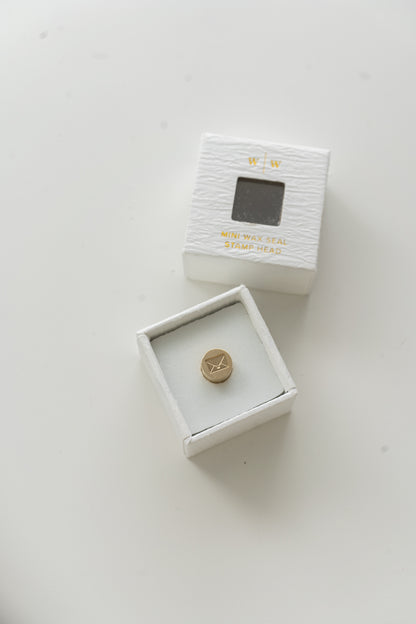 ​A mini brass wax seal stamp head with an envelope design in a textured white ring-style box, with its lid beside it