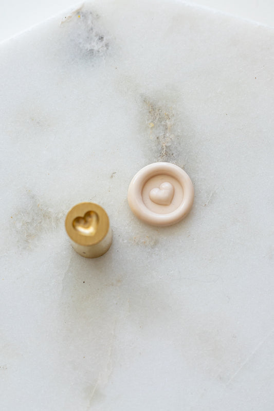 A mini brass wax seal stamp head with a 3D heart design beside a blush wax seal impression on a white marble surface