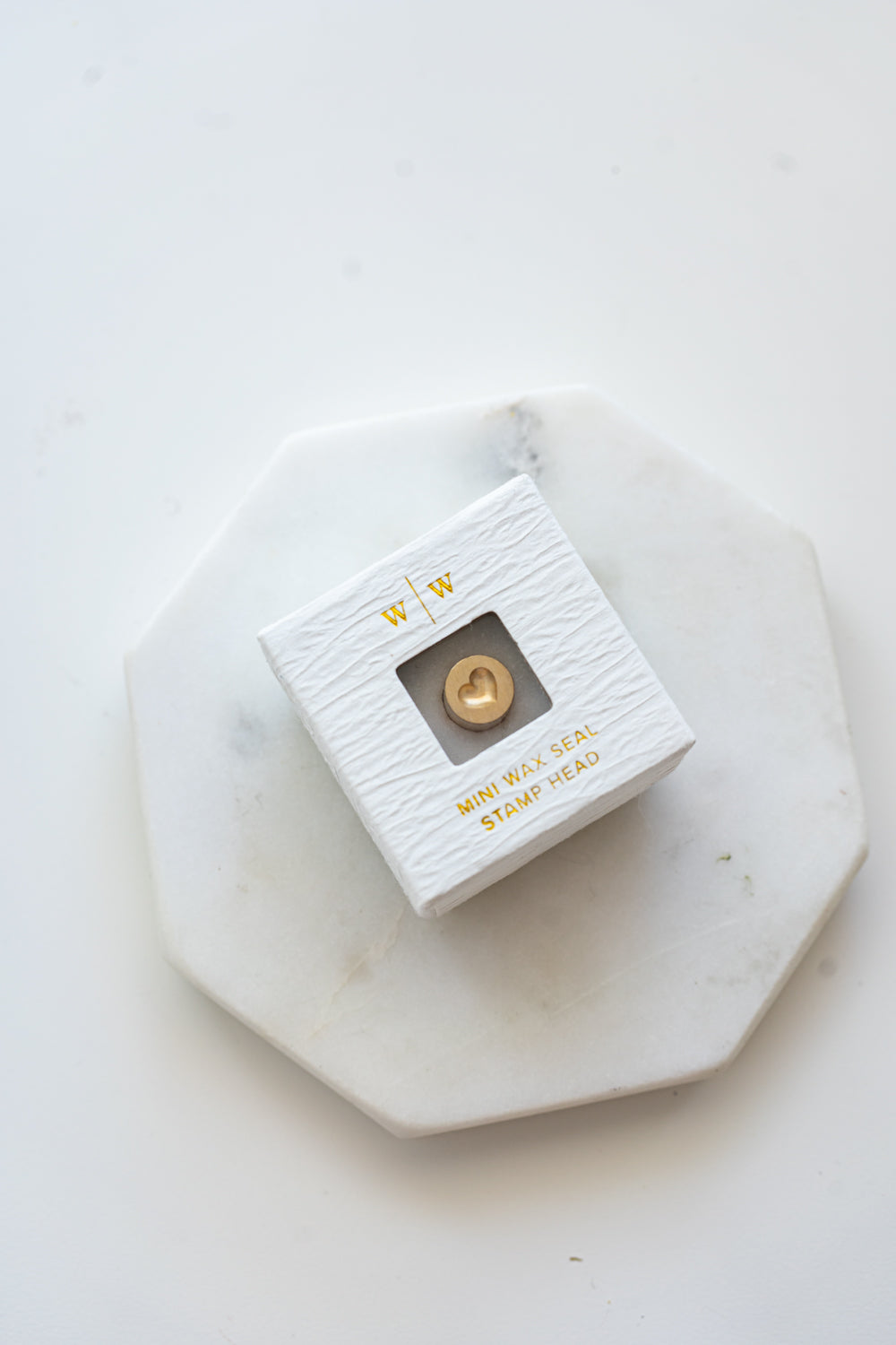 A mini brass wax seal stamp head with a 3D heart design in a textured white ring-style box on a marble coaster