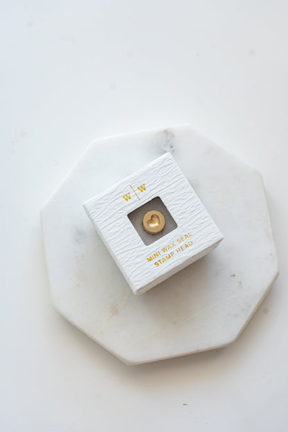A mini brass wax seal stamp head with a 3D heart design in a textured white ring-style box on a marble coaster