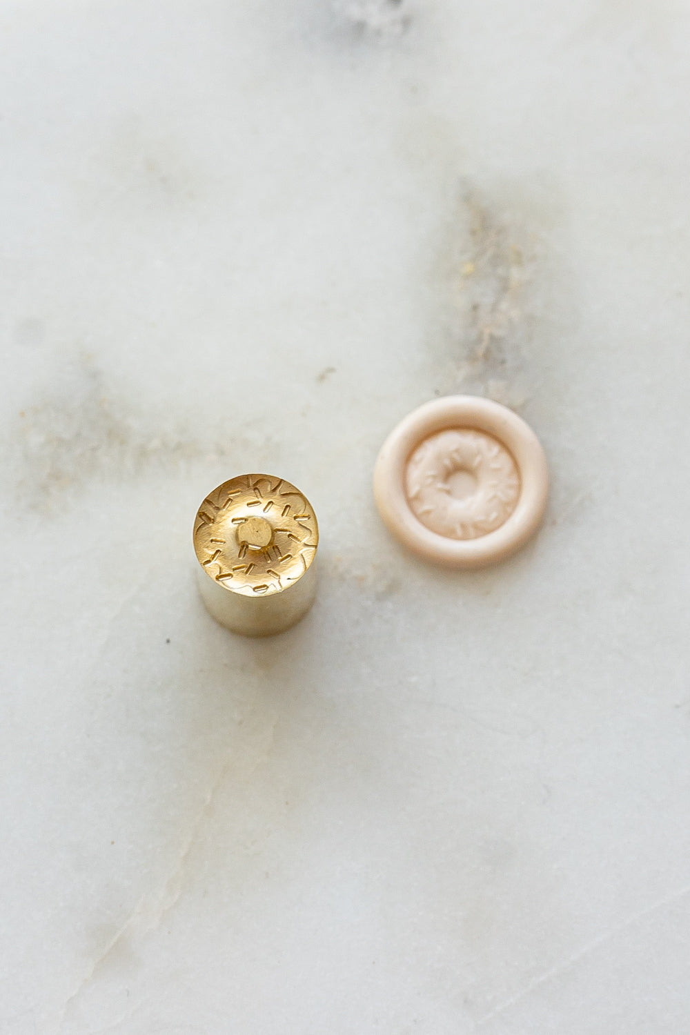 ​A mini brass wax seal stamp head with a 3D donut design beside a blush wax seal impression on a white marble surface