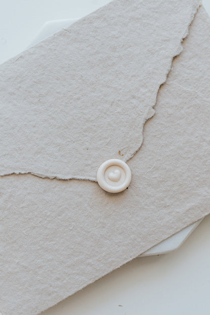 A mini 9mm blush wax seal with a 3D heart design on a textured taupe envelope