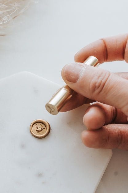Hand holding brass mini wax seal stamp head with dove design, pressed into gold wax on marble surface