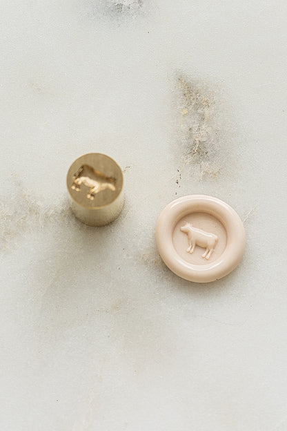 A mini brass wax seal stamp head with a 3D cow design beside a blush wax seal impression on a white marble surface