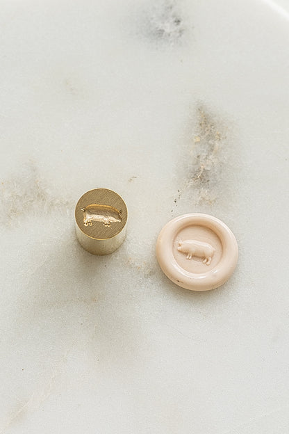 A mini brass wax seal stamp head with a 3D pig design beside a blush wax seal impression on a white marble surface