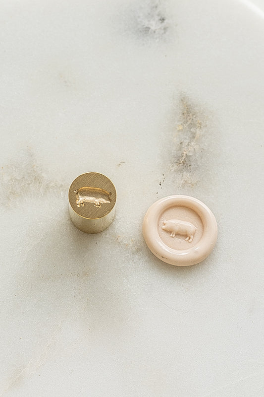 ​A mini brass wax seal stamp head with a 3D pig design beside a blush wax seal impression on a white marble surface