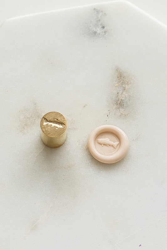 ​A mini brass wax seal stamp head with a 3D fish design beside a blush wax seal impression on a white marble surface