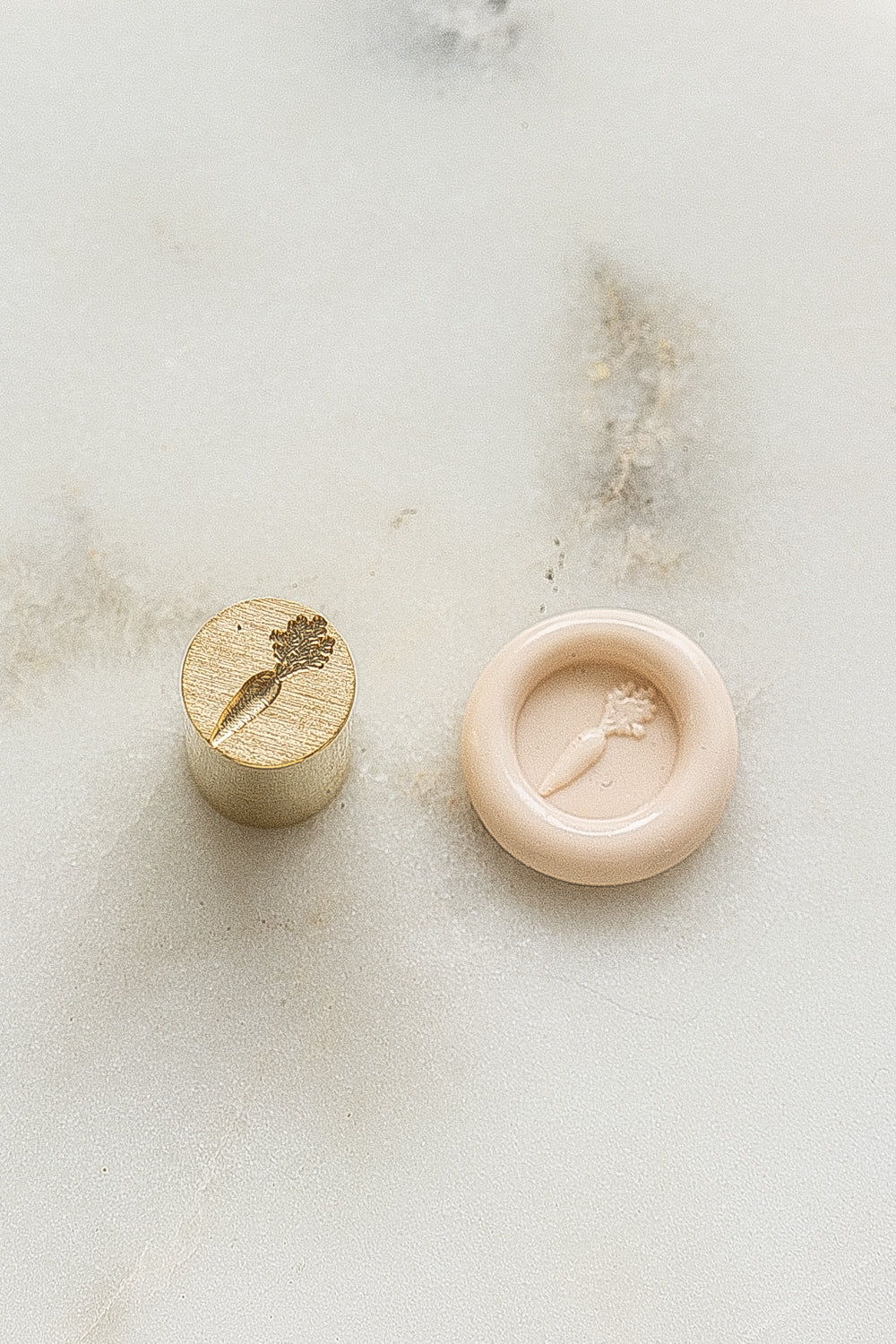 ​A mini brass wax seal stamp head with a 3D carrot design beside a blush wax seal impression on a white marble surface