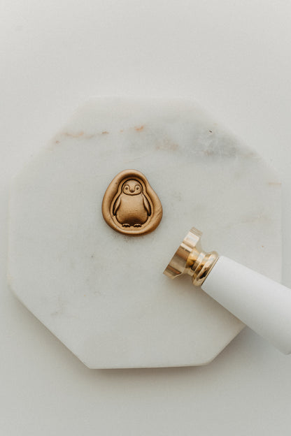 Gold penguin wax seal and white-handled brass wax seal stamp on a white marble tile.
