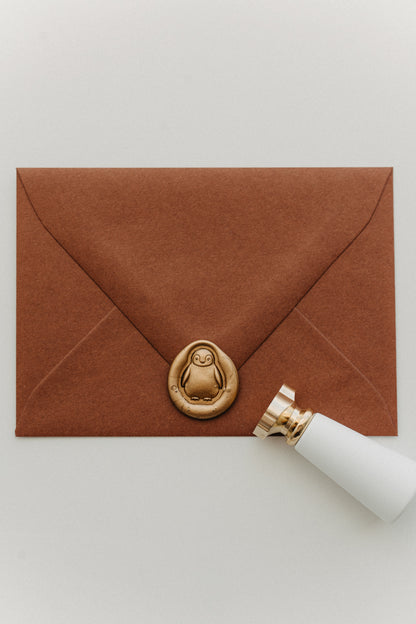 Gold penguin wax seal on a warm brown envelope beside a white-handled brass wax seal stamp.