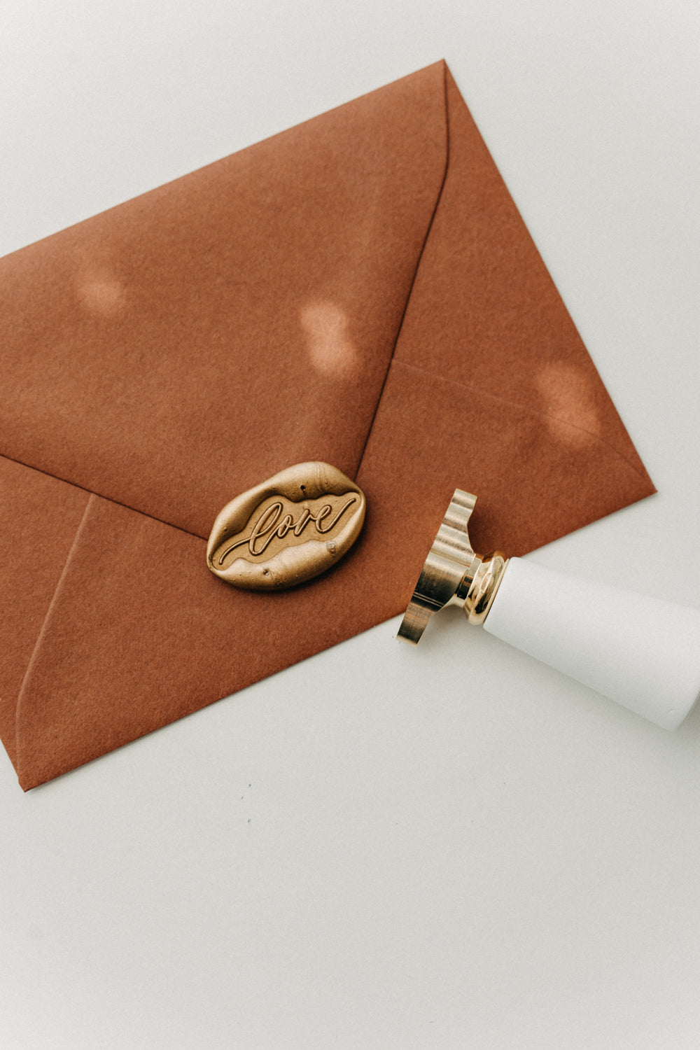 Gold “love” wax seal on a warm brown envelope beside a white-handled brass wax seal stamp.