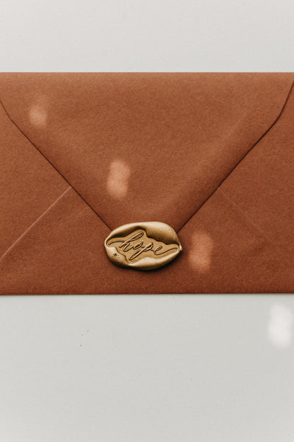 Metallic gold “hope” wax seal securing a warm brown envelope, close-up stationery detail.