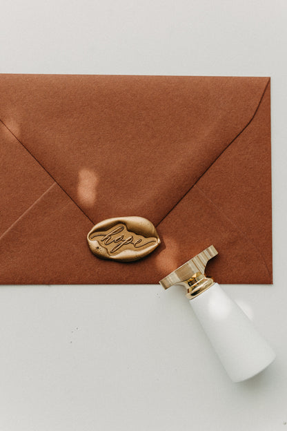 Gold “hope” wax seal on a warm brown envelope beside a white-handled brass wax seal stamp.