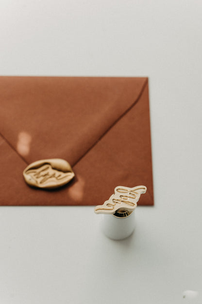 White-handled brass “hope” wax seal stamp displayed with a gold wax-sealed warm brown envelope.