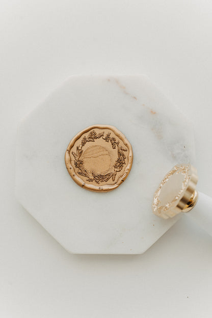 Metallic gold wax seal embossed with a botanical wreath design beside a white-handled brass wax stamp on white marble.