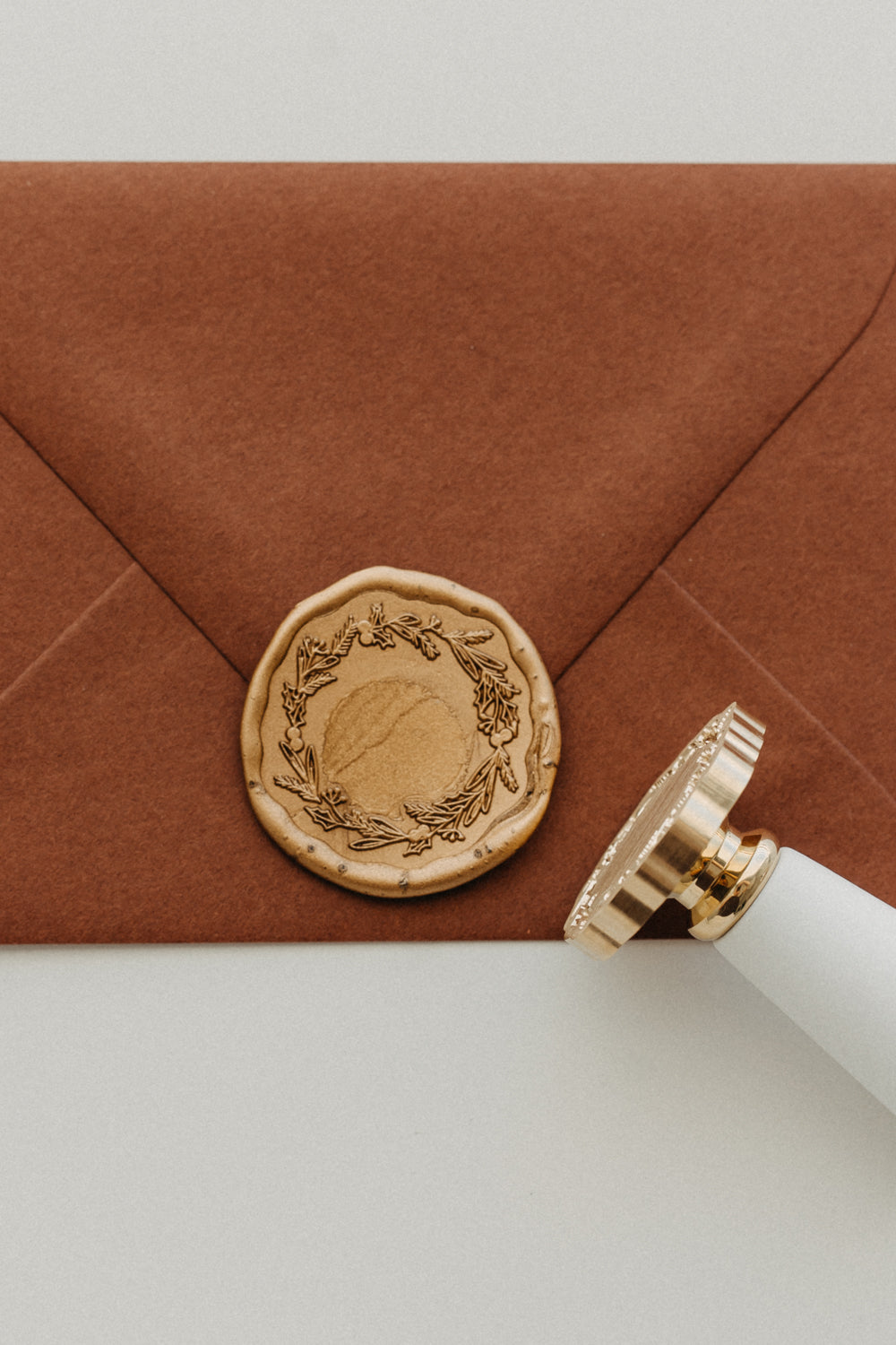 Gold botanical wreath wax seal securing a warm brown envelope beside a white-handled brass wax seal stamp.