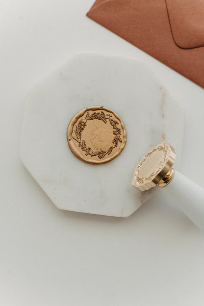 Metallic gold botanical wreath wax seal and white-handled brass wax stamp displayed on a white marble tile.