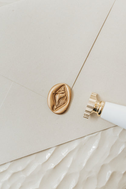 Gold conch shell wax seal on a cream envelope beside a brass wax seal stamp for sealing wedding invitations and letters.