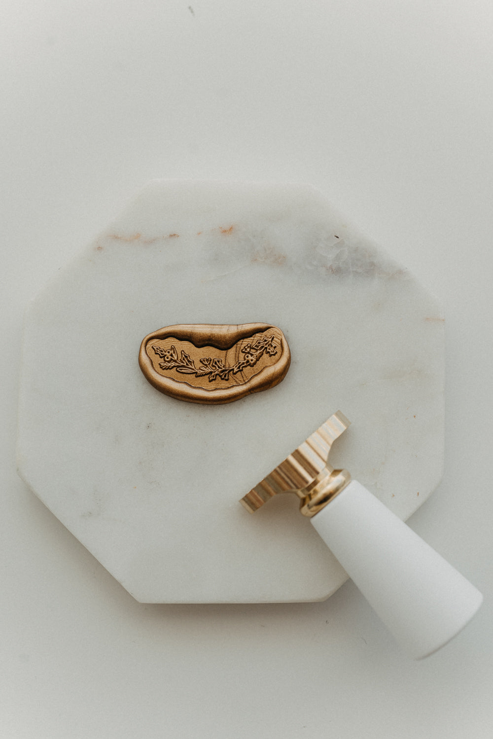 Gold botanical garland wax seal and white-handled brass wax seal stamp displayed on a white marble tile.