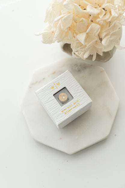 Mini wax seal stamp head in white textured box with gold foil text, styled with dried flowers on marble tray