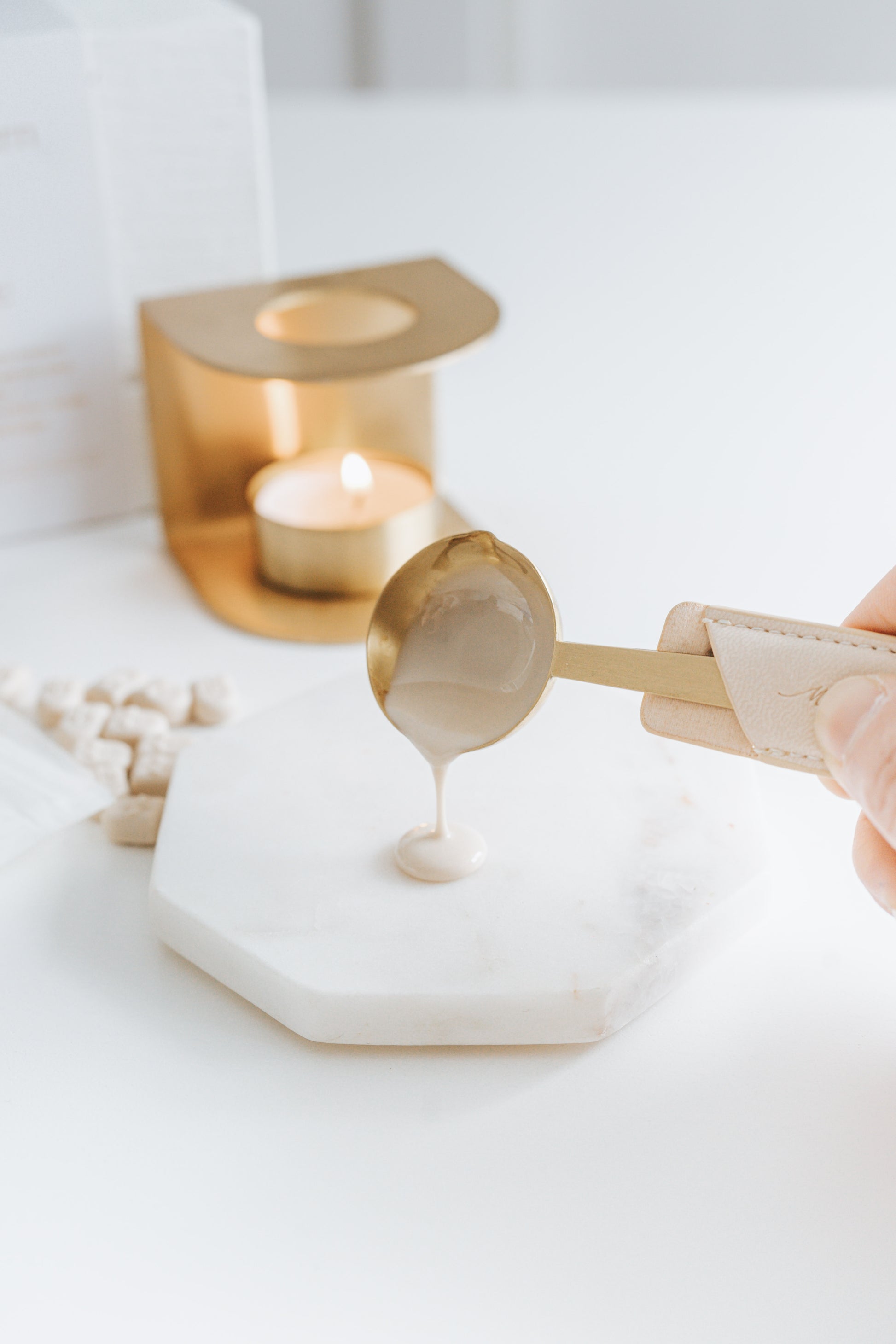 A hand pours melted beige wax from a brass spoon onto a marble slab. A brass wax seal stove with a tea light is visible in the background.