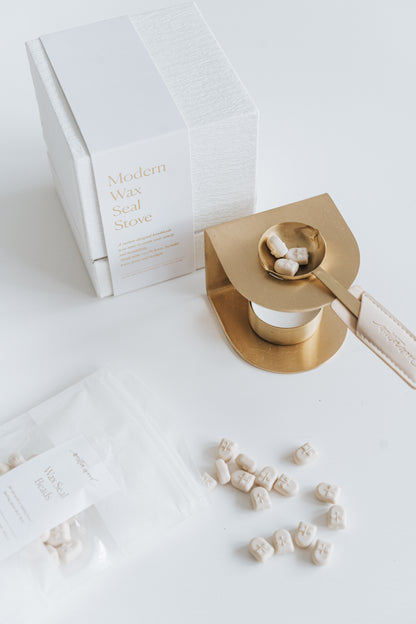 Brass wax seal stove with a melting spoon holding ivory wax beads, placed beside its textured white packaging and a scattered pouch of wax beads
