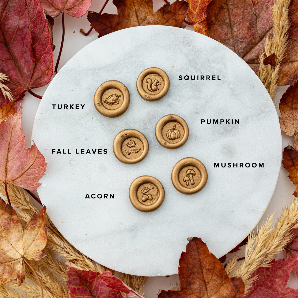 Flat lay of gold wax seal impressions featuring fall-themed designs—turkey, squirrel, pumpkin, fall leaves, acorn, and mushroom—on a marble surface surrounded by autumn leaves and wheat stalks.