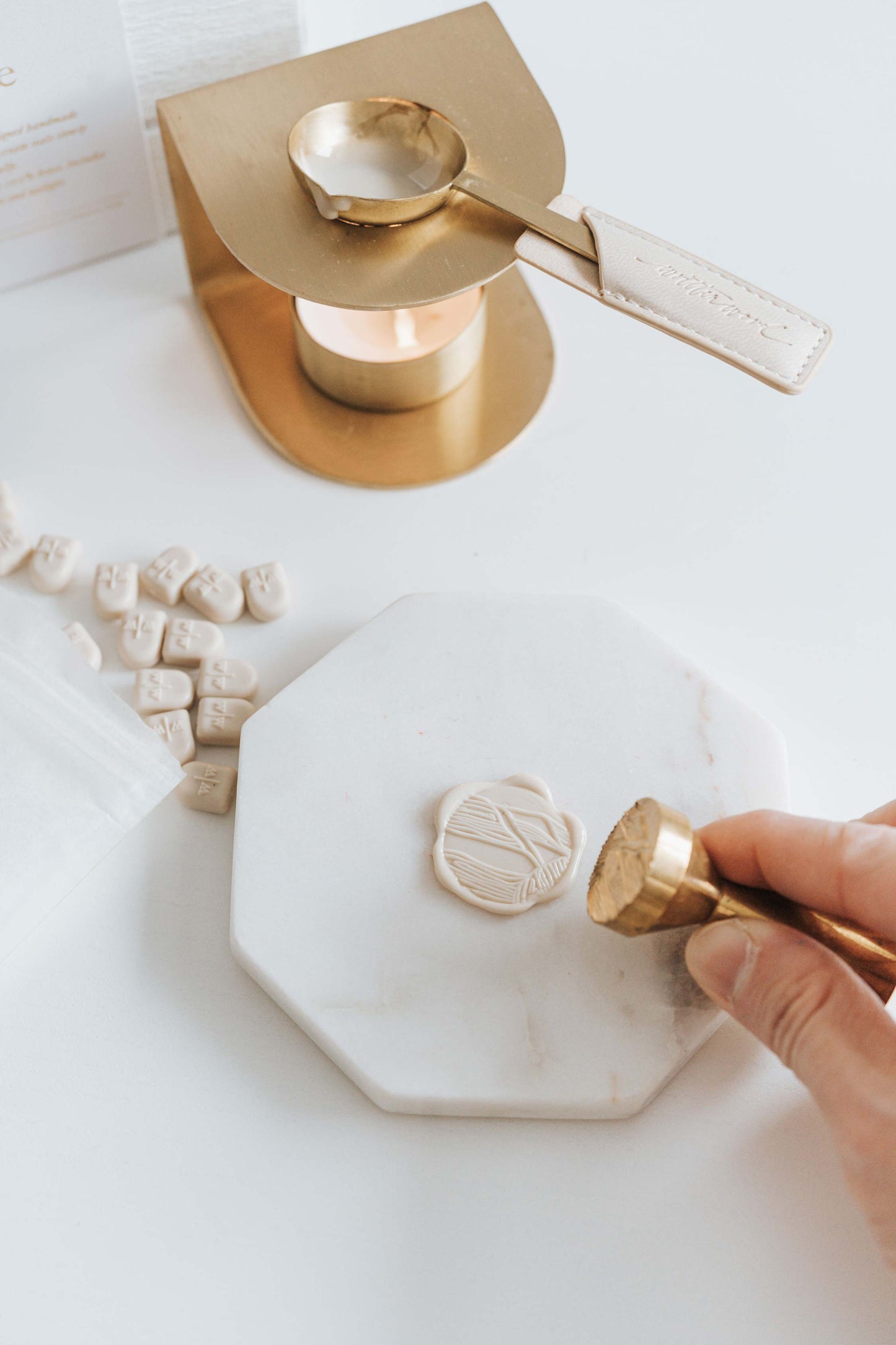 Brass wax seal stove with a tea light and wax spoon, melting beige wax beads. A hand and presses a brass stamp onto a wax seal on a marble slab