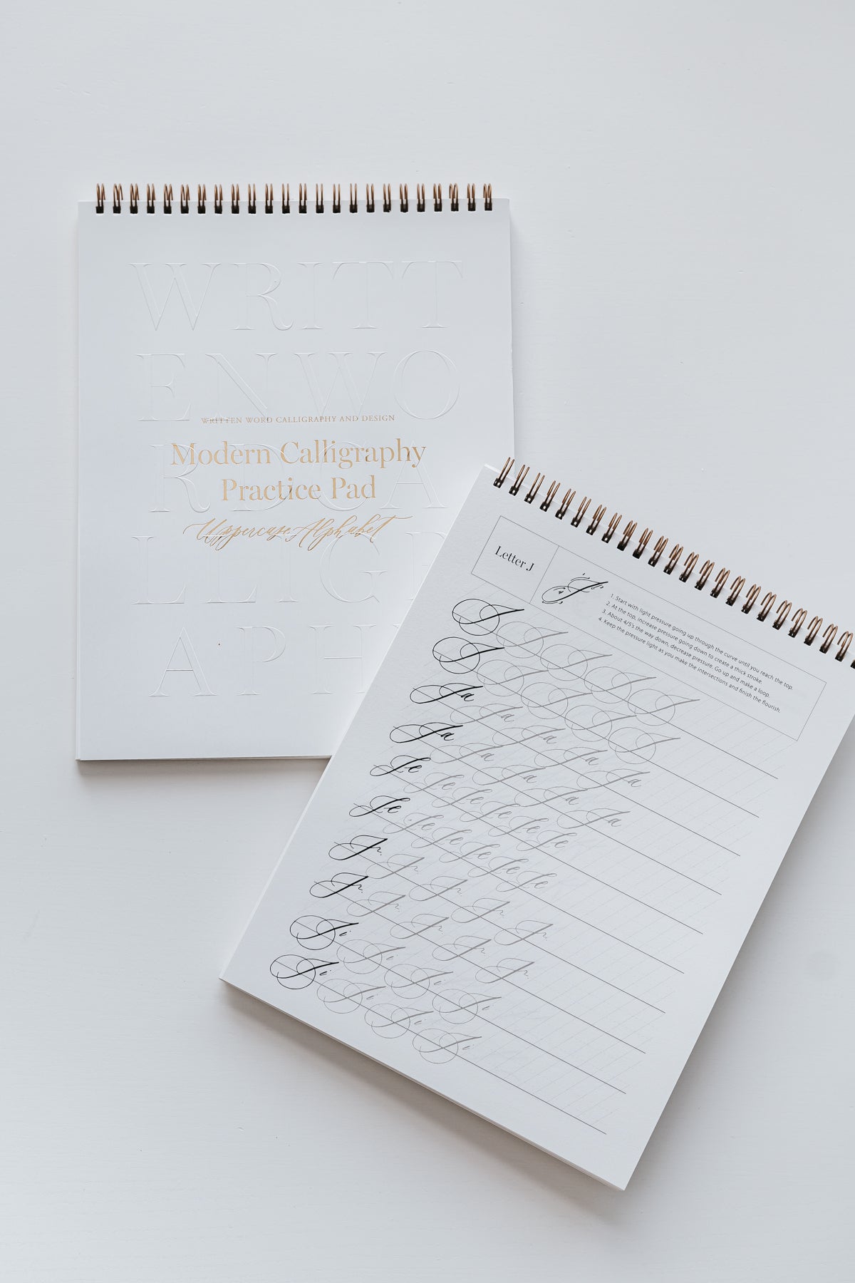 Two modern calligraphy practice pads with letters and examples for beginners, with spiral binding and gold accents