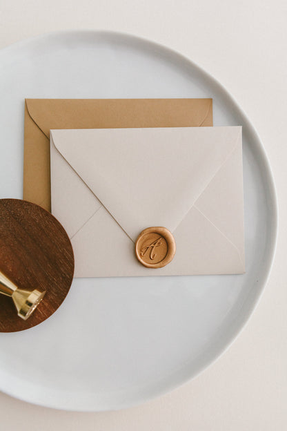 Brass wax seal stamp on a wooden dish beside envelopes with a gold A Monogram wax seal, styled on a white ceramic plate.