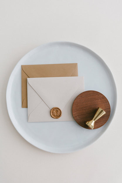 Brass wax seal stamp on a wooden dish beside envelopes with a gold floral crest wax seal, styled on a white ceramic plate.