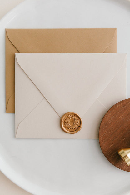 Brass wax seal stamp on a wooden dish beside envelopes with a gold floral crest wax seal, styled on a white ceramic plate.