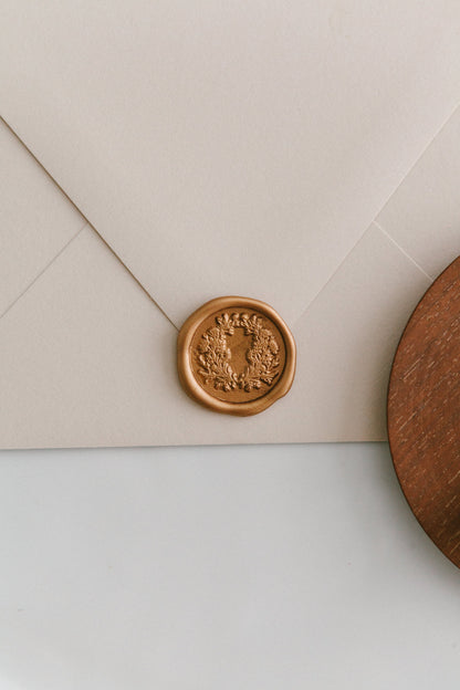 A golden floral crest wax seal, styled on a beige sealed envelope and a wooden dish on the side