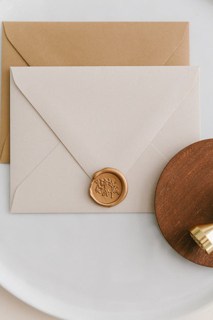 Brass wax seal stamp on a wooden dish beside envelopes with a gold Jasmine Vine wax seal, styled on a white ceramic plate.