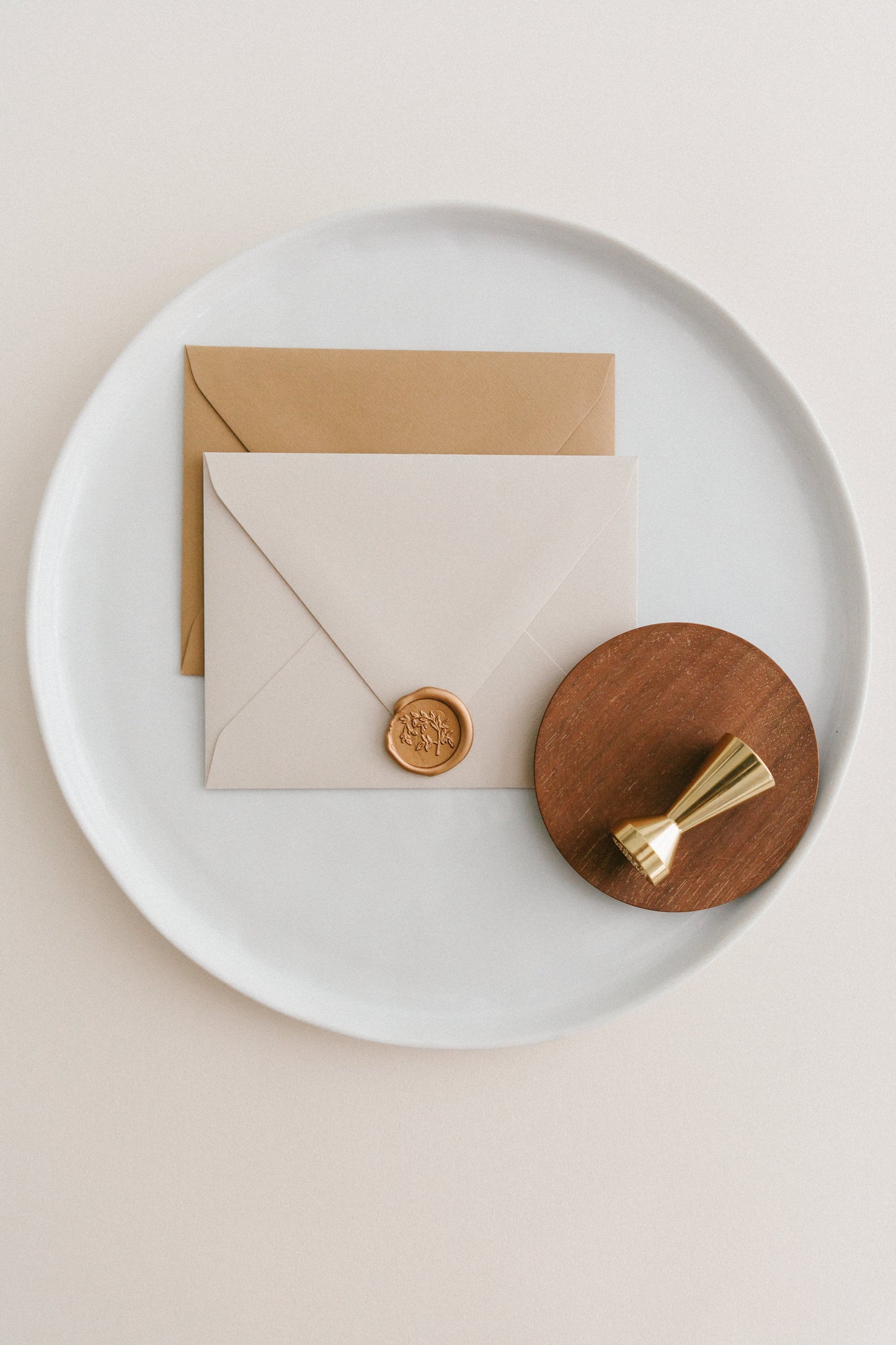 Brass wax seal stamp on a wooden dish beside envelopes with a gold Jasmine Vine wax seal, styled on a white ceramic plate.