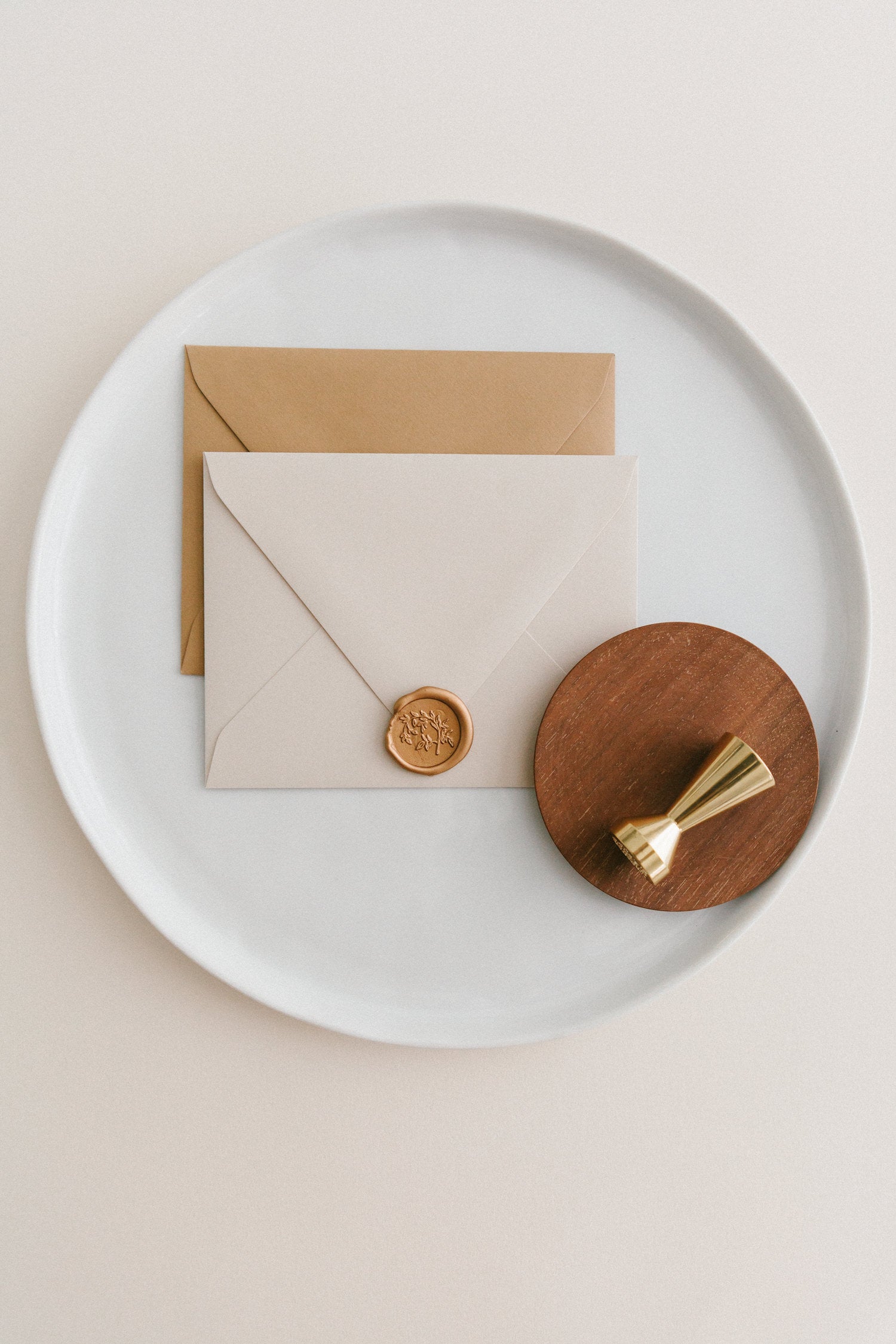 Brass wax seal stamp on a wooden dish beside envelopes with a gold Jasmine Vine wax seal, styled on a white ceramic plate.